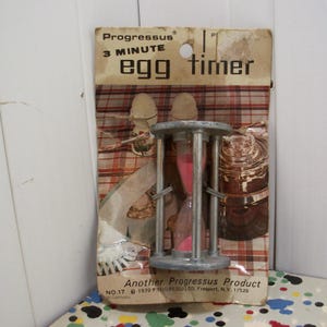 May include: Vintage Progressus 3-minute egg timer in original packaging. The timer has a silver metal frame with pink sand inside. The packaging is a worn cardboard with the text "Progressus 3 MINUTE egg timer".