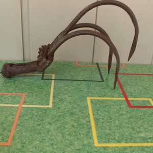 May include: An antique, rusty metal hay hook with four curved tines. The tool has a handle and a claw-like design, likely used for agricultural purposes. The hook is set against a green surface with colorful square patterns.