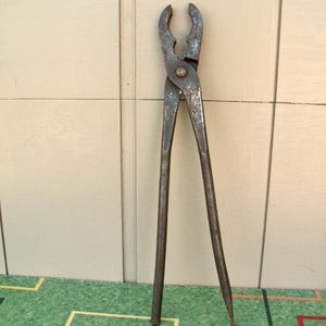 May include: Vintage metal pliers with long handles and a curved, toothed head. The tool has a worn, rusty appearance, suggesting age and use. The pliers are designed for gripping and manipulating objects.