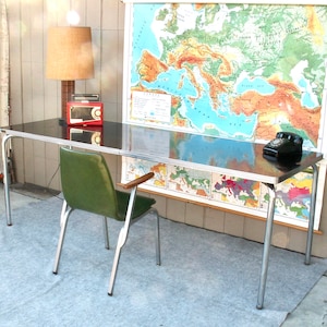 May include: A black and silver metal desk with a green chair. The desk has a black top and silver legs. The chair has a green upholstered seat and a silver frame. There is a red radio and a black rotary phone on the desk. A map of Europe is hanging on the wall behind the desk.