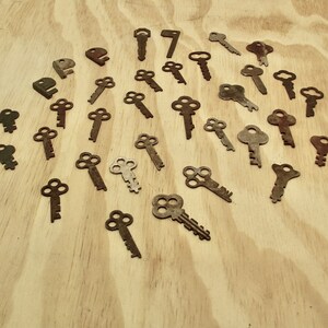May include: A collection of vintage keys with various designs and finishes, including brass, copper, and nickel. The keys are arranged on a light wood surface.