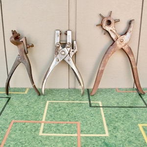 May include: Three vintage metal tools, including a leather punch, a rivet setter, and a revolving punch plier. The tools are made of metal and show signs of wear, with a rustic, aged appearance. The tools are arranged on a green surface.