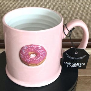 May include: Pink ceramic mug with a glazed finish, featuring a hand-painted donut design with pink frosting and sprinkles. The mug has a large handle and a tag that reads "HAND CRAFTED & PAINTED".