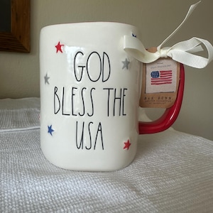 May include: White ceramic mug with a red handle and rim, decorated with red, white, and blue stars. The mug features the text "GOD BLESS THE USA" in black lettering. A tag with an American flag is attached with a white ribbon.