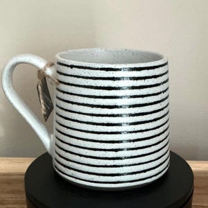 Peut inclure: Mug en céramique blanche avec des rayures horizontales noires. La tasse a une anse incurvée et un intérieur moucheté. Une petite étiquette est attachée à l'anse avec de la ficelle. La tasse est posée sur une surface noire.