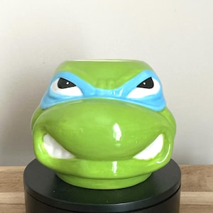 Pode incluir: Uma caneca de cerâmica em forma da cabeça de Leonardo das Tartarugas Ninja. A caneca é verde com uma máscara azul, dentes brancos e olhos pretos. A caneca está sobre uma superfície preta.