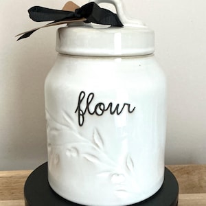 Può includere: Barattolo di farina in ceramica bianca con coperchio e manico. La parola "flour" è scritta in nero sul davanti. Un nastro nero e un'etichetta sono legati al manico. Il barattolo ha un design floreale.