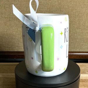 May include: White ceramic mug with a green handle and the words "READ LOVE" in black. The mug is decorated with colorful star shapes and a light blue ribbon tied around the handle. A blue tag is attached to the ribbon.