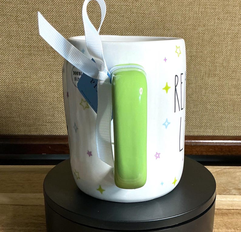 May include: White ceramic mug with a green handle and the words "READ LOVE" in black. The mug is decorated with colorful star shapes and a light blue ribbon tied around the handle. A blue tag is attached to the ribbon.