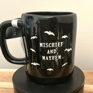 Puede incluir: Taza de cer&aacute;mica negra con siluetas de murci&eacute;lagos blancos y las palabras "MISCHIEF AND MAYHEM". La taza tiene un asa grande y una etiqueta adjunta. Una taza con tem&aacute;tica de Halloween.