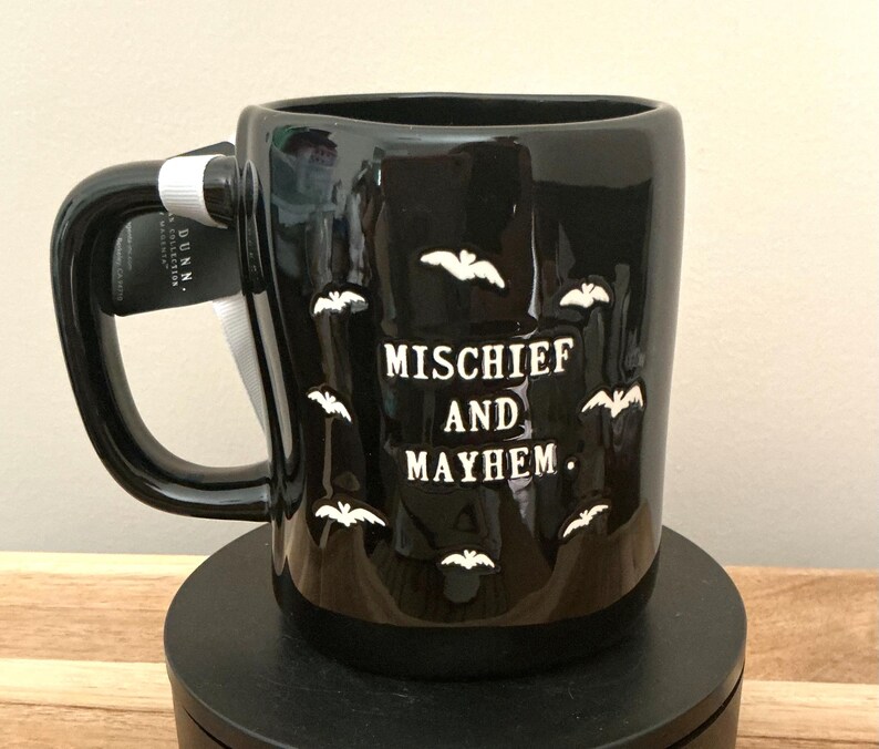 Puede incluir: Taza de cer&aacute;mica negra con siluetas de murci&eacute;lagos blancos y las palabras "MISCHIEF AND MAYHEM". La taza tiene un asa grande y una etiqueta adjunta. Una taza con tem&aacute;tica de Halloween.