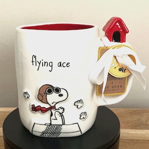 May include: White ceramic mug featuring Snoopy as the Flying Ace, with a red interior. The mug has a red doghouse accent on the handle and the words "flying ace" printed on the side. A tag with a white ribbon is attached to the handle.