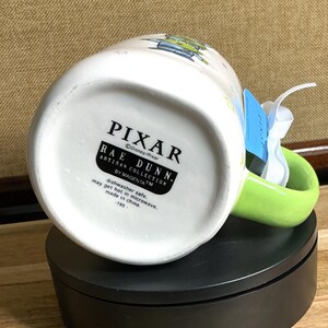 May include: White ceramic mug with a green handle, featuring a Pixar design. The mug has the Pixar and Rae Dunn logos. The bottom of the mug has text that reads "dishwasher safe" and "made in china."