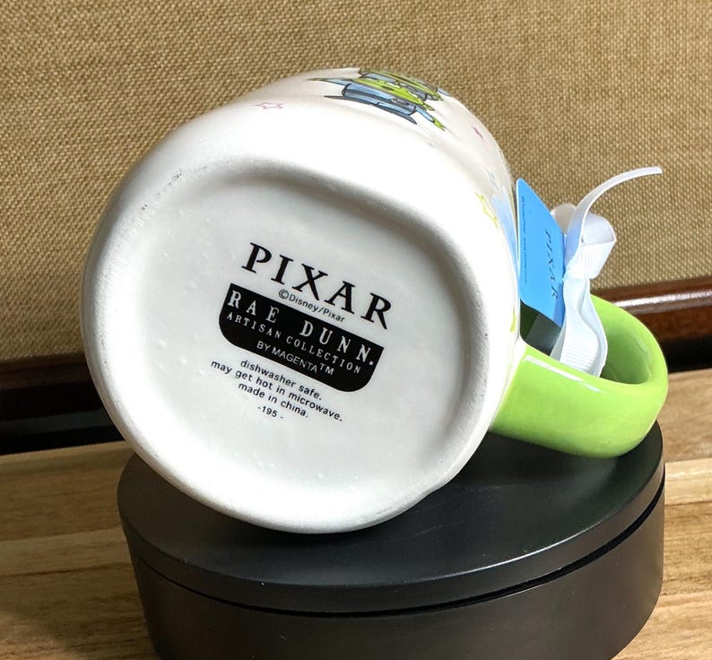 May include: White ceramic mug with a green handle, featuring a Pixar design. The mug has the Pixar and Rae Dunn logos. The bottom of the mug has text that reads "dishwasher safe" and "made in china."