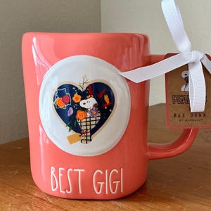 Könnte beinhalten: Korallenfarbene Keramik-Tasse mit einem weißen herzförmigen Aufdruck mit Snoopy in einem Blumenkorb. Die Tasse trägt den Text "BEST GIGI" und ein weißes Band, das an einem Anhänger mit der Aufschrift "PEANUTS" befestigt ist.