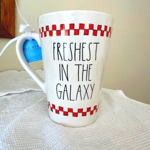 May include: A white ceramic mug featuring a red checkered pattern at the top and bottom rims. The text "FRESHEST IN THE GALAXY" is printed in black lettering on the front. A blue product tag is attached to the handle with a white ribbon.