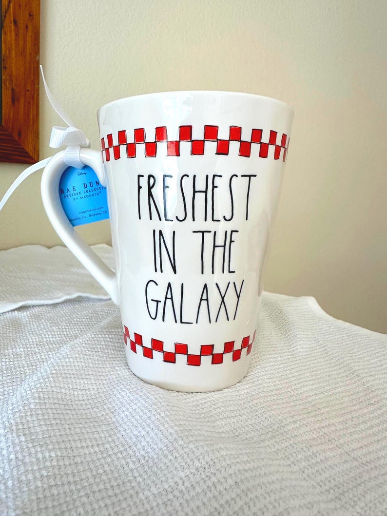 May include: A white ceramic mug featuring a red checkered pattern at the top and bottom rims. The text "FRESHEST IN THE GALAXY" is printed in black lettering on the front. A blue product tag is attached to the handle with a white ribbon.