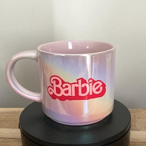 Puede incluir: Una taza de café rosa nacarado con la palabra "Barbie" en escritura roja. La taza tiene un asa curva y un acabado brillante, que refleja un arcoíris de colores. La taza está sobre una superficie negra.