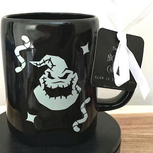 Puede incluir: Taza de cerámica negra con un diseño de Oogie Boogie que brilla en la oscuridad de The Nightmare Before Christmas. La taza tiene un asa negra y una cinta blanca atada a una etiqueta negra.