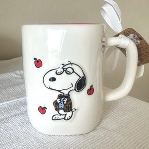 May include: White ceramic mug featuring a cartoon Snoopy character wearing glasses and a jacket, with red apple illustrations. The mug has a white handle and a red interior. A white ribbon and a tag are attached to the handle.