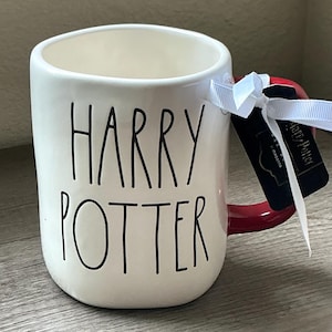 May include: A cream-colored ceramic mug with the text HARRY POTTER printed in black, sans-serif lettering. The mug features a dark red handle and a black rectangular product tag tied to the handle with a thin white ribbon.