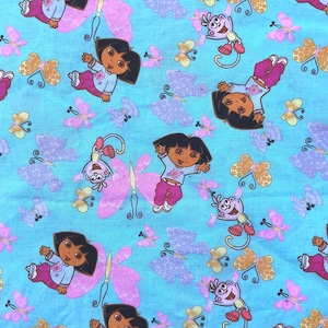 May include: A light blue fabric featuring a repeating pattern of Dora the Explorer characters wearing light blue shirts and pink pants, alongside purple and gold butterflies. The characters are depicted in various orientations across the surface.