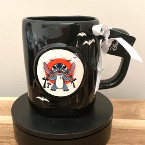Puede incluir: Taza de cer&aacute;mica negra con un colorido gr&aacute;fico de Stitch en un c&iacute;rculo blanco con fondo rojo. La taza tiene un asa negra y est&aacute; decorada con siluetas de murci&eacute;lagos. Se adjuntan una cinta blanca y una etiqueta de Disney.
