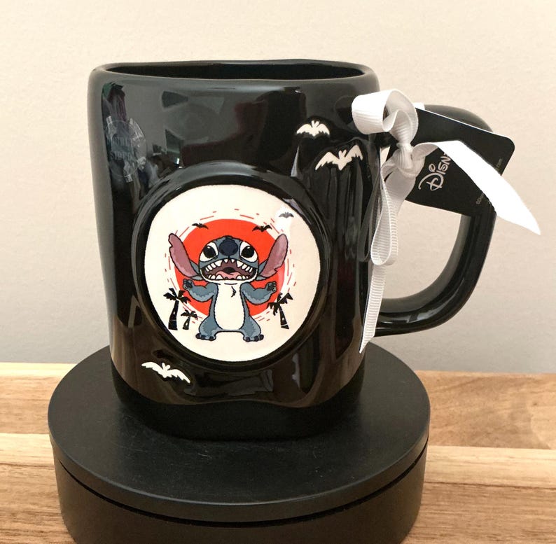 Puede incluir: Taza de cer&aacute;mica negra con un colorido gr&aacute;fico de Stitch en un c&iacute;rculo blanco con fondo rojo. La taza tiene un asa negra y est&aacute; decorada con siluetas de murci&eacute;lagos. Se adjuntan una cinta blanca y una etiqueta de Disney.