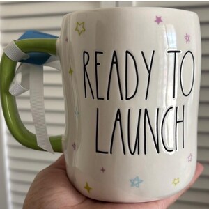 May include: A white ceramic mug with a black handle and the text "But first, coffee" in a handwritten style. The mug is on a white surface. The mug is 4 inches tall.