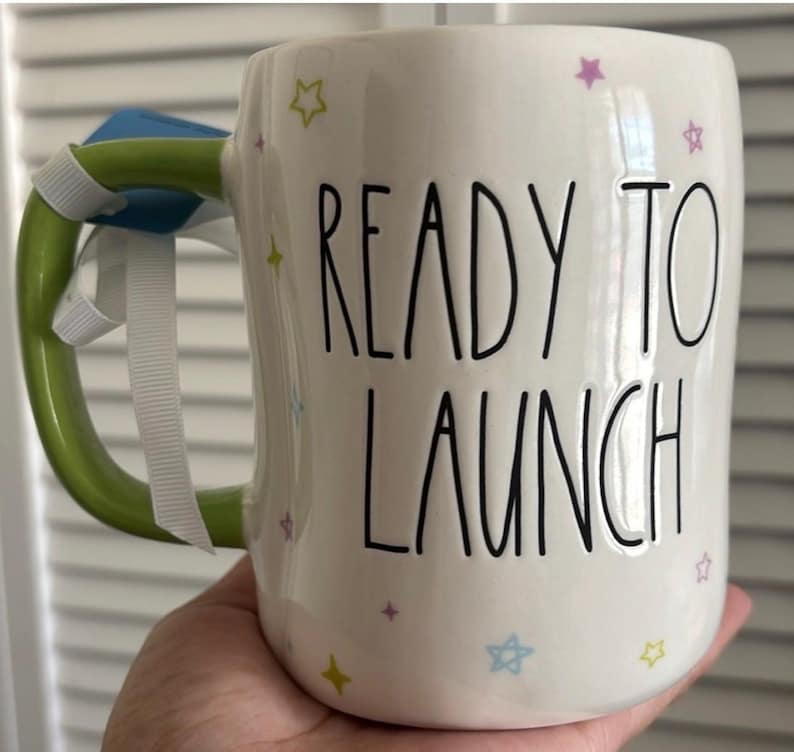 May include: A white ceramic mug with a black handle and the text "But first, coffee" in a handwritten style. The mug is on a white surface. The mug is 4 inches tall.