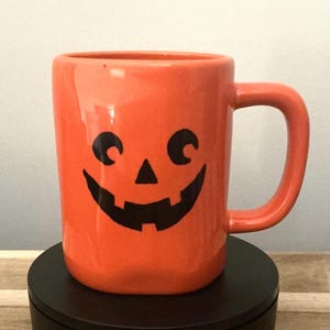 Puede incluir: Taza de cerámica naranja con una cara de calabaza negra. La taza tiene un asa y está sobre una superficie negra. Un artículo con temática de Halloween.