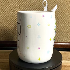 May include: A white ceramic mug with a black handle and the text "But First, Coffee" in a black, handwritten-style font. The mug is on a white surface.