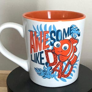 Peut inclure: Mug en céramique blanche avec intérieur et anse orange. Le mug présente le texte "AWESOME LIKE DAD" en bleu et orange, ainsi qu'un poisson orange et blanc de dessin animé et des accents de feuilles bleues.