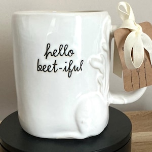 May include: White ceramic mug with the text "hello beet-iful" in black script. The mug has a curved handle and embossed floral details. A cream-coloured ribbon and tag are attached to the handle.