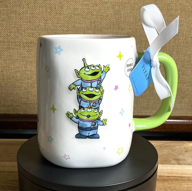 May include: White ceramic mug featuring a stack of three green alien characters from the Pixar movie Toy Story. The mug has a green handle and is decorated with colorful stars. A blue tag with the word "PIXAR" is attached with a white ribbon.
