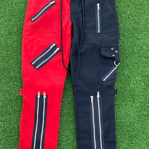 May include: Black and red pants with multiple zipper details. The pants have a cargo pocket on the right leg and a zipper detail on the left leg. The pants are made of a cotton blend.