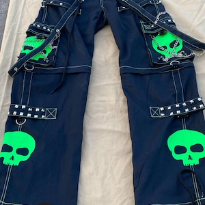 May include: Black cargo pants with green skull graphics and neon green stitching. The pants have multiple straps and buckles, and a chain detail.