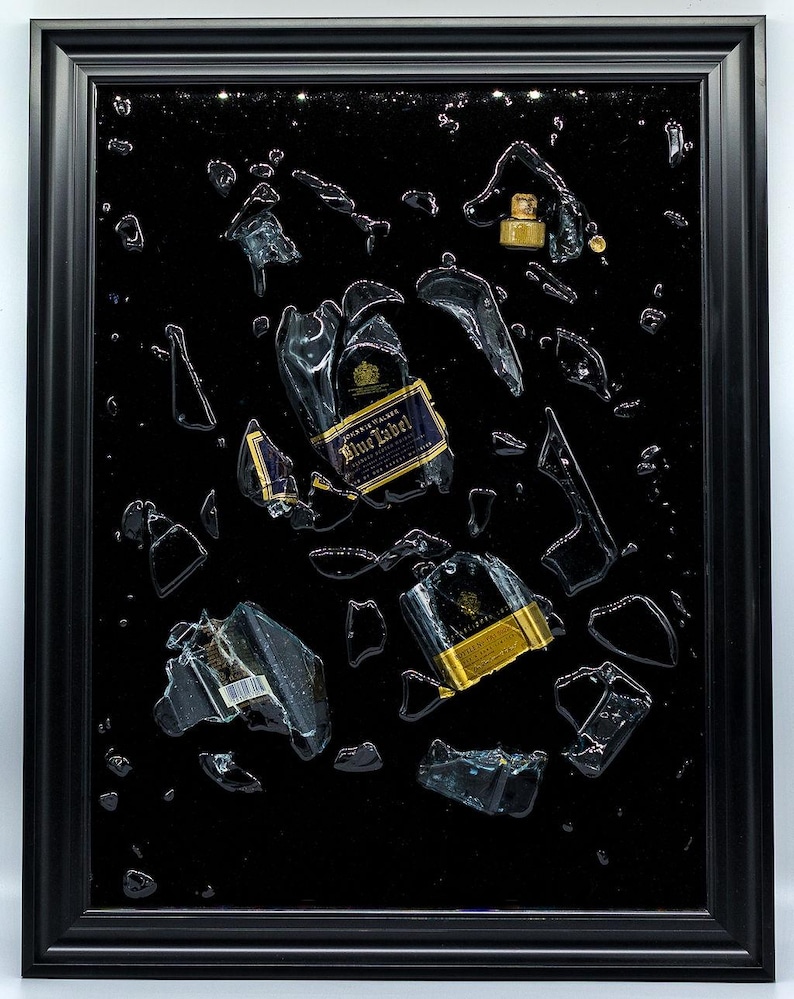 May include: A framed artwork featuring shattered glass and liquid droplets against a black background. Fragments of a bottle with a blue and gold label are scattered throughout. The artwork is encased in a black frame.