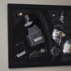 Może przedstawiać: Oprawiona grafika przedstawiająca fragmenty butelek po alkoholu, w tym Woodford Reserve i Buffalo Trace. Przezroczyste szklane elementy ułożone są na czarnym tle w czarnej ramie, tworząc unikalną ekspozycję.