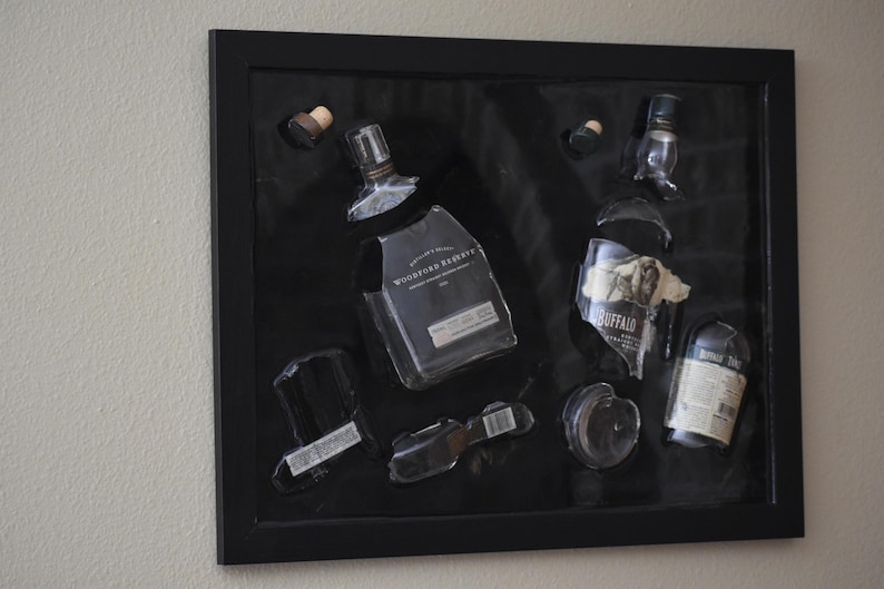 Może przedstawiać: Oprawiona grafika przedstawiająca fragmenty butelek po alkoholu, w tym Woodford Reserve i Buffalo Trace. Przezroczyste szklane elementy ułożone są na czarnym tle w czarnej ramie, tworząc unikalną ekspozycję.