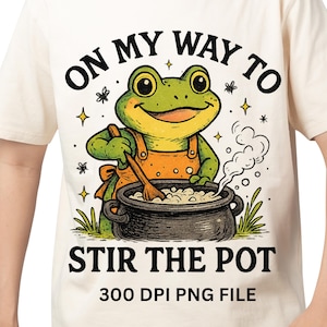 May include: Cream-colored t-shirt featuring a cartoon frog wearing an orange apron, stirring a pot. The text "ON MY WAY TO STIR THE POT" is above and below the frog. Additional text reads "300 DPI PNG FILE".
