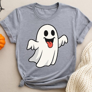 Puede incluir: Camiseta gris claro con un fantasma de dibujos animados sonriendo, con ojos negros y lengua roja. Una pequeña calabaza naranja y una telaraña negra completan la composición, evocando Halloween.