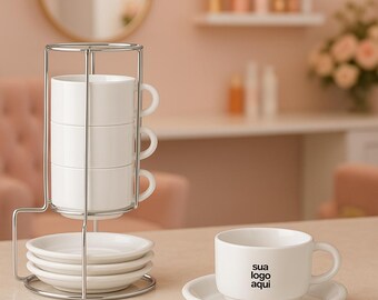 May include: A white ceramic espresso cup and saucer set with the text "sua logo aqui" printed on the cup. The set includes a stack of three cups and saucers in a chrome metal stand. The background is a soft pink.