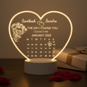 May include: A heart-shaped LED night light with the names "Santhosh" and "Sandra" and the text "THE DAY I FOUND YOU I found love JANUARY 2025". The light has a calendar with the date highlighted. A bouquet of flowers is on the left.