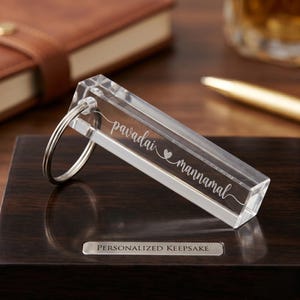 May include: A clear rectangular acrylic keychain featuring the engraved text pavadai heart mannamal. The keychain includes a silver metal split ring attached to one end, showcasing a transparent prism-like design with smooth edges and a polished finish.