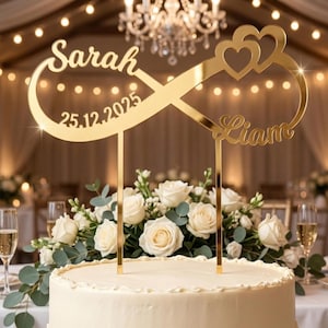 Peut inclure: Un cake topper doré avec les noms "Sarah" et "Liam" et la date "25.12.2025". Le topper présente un symbole infini avec deux cœurs. Le gâteau est décoré de roses blanches et de verdure.