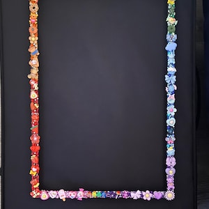 May include: A rectangular picture frame featuring a vibrant, rainbow-colored border decorated with an assortment of small, colorful plastic charms including flowers, hearts, stars, food items, and animals arranged in a spectrum of colors.
