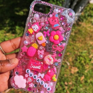 May include: A clear phone case with a pink, glittery design. The case is decorated with various charms, including a pineapple, a donut, a flower, a bear, and a cat. The text "Sweet Eurasia" is spelled out in pink letters on the case.