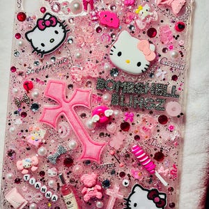 May include: A pink and clear phone case decorated with various charms, including Hello Kitty faces, bows, a cross, and the text "Girl Boss" and "Bombshell Illingz". The case is covered in glitter and small beads.