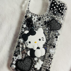 May include: A clear phone case decorated with black and white rhinestones, a Hello Kitty character, a black bow, and the word "Scorpio" with a letter K in the middle.